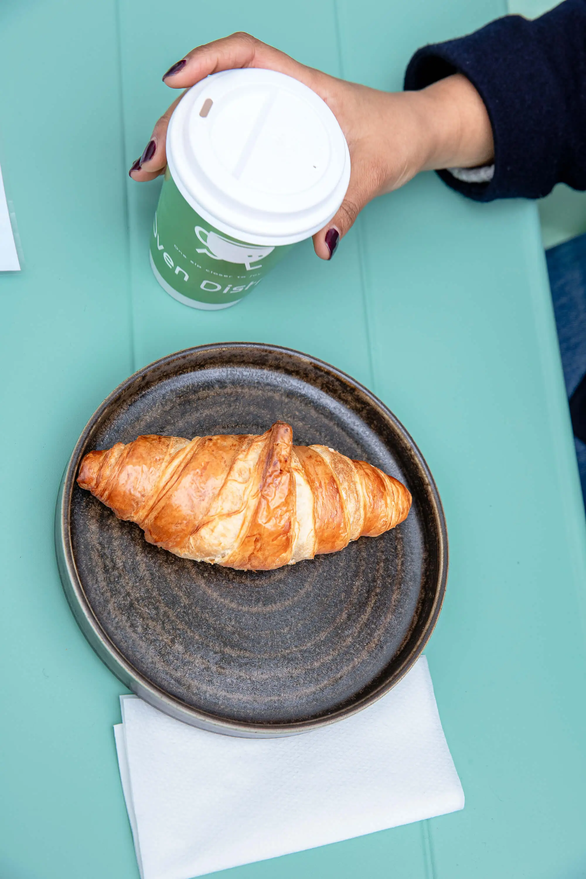 Hand holding a green coffee cup above a dark plate with a croissant on a teal table.