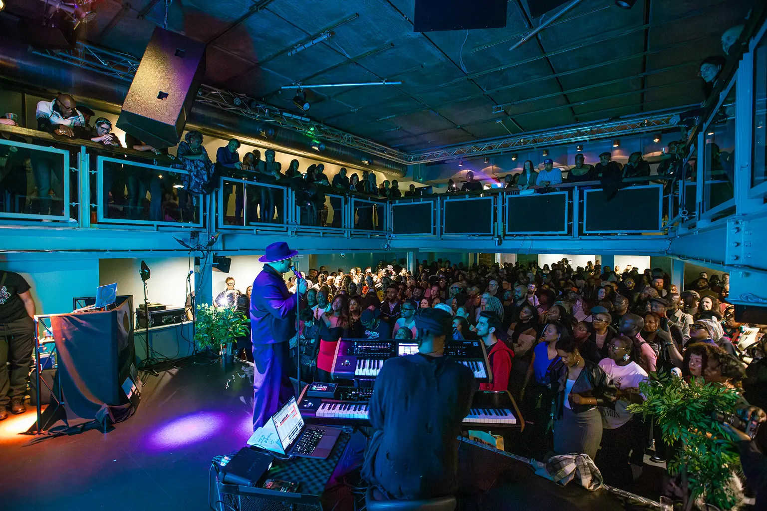 Musician in purple suit and hat performs on stage with keyboard player for a packed crowd in a two-level venue.