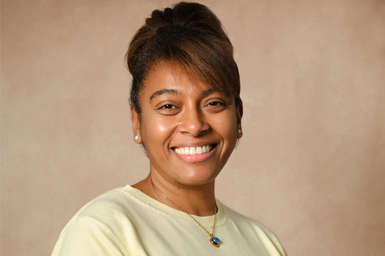 Smiling woman with pearl earrings and necklace wearing a light yellow top against a beige background.