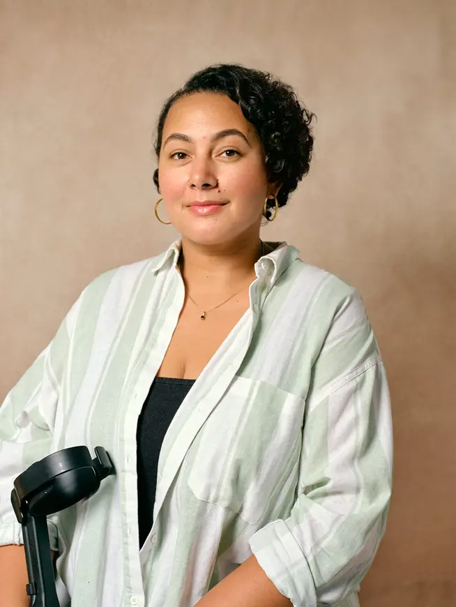 Woman with short curly hair wearing gold hoop earrings and a striped shirt stands holding a black object.