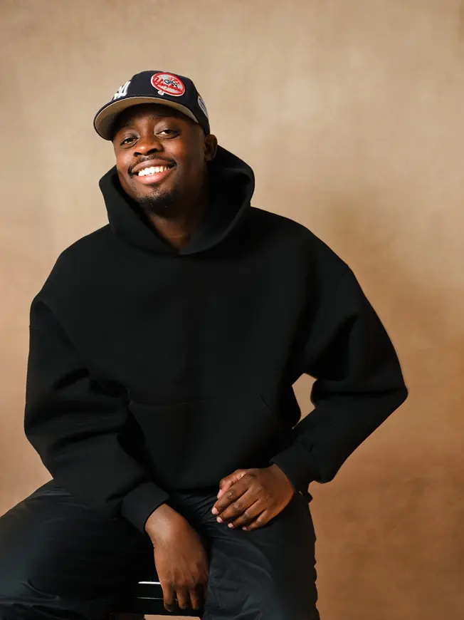 Smiling man in black hoodie and baseball cap sitting with neutral brown background.