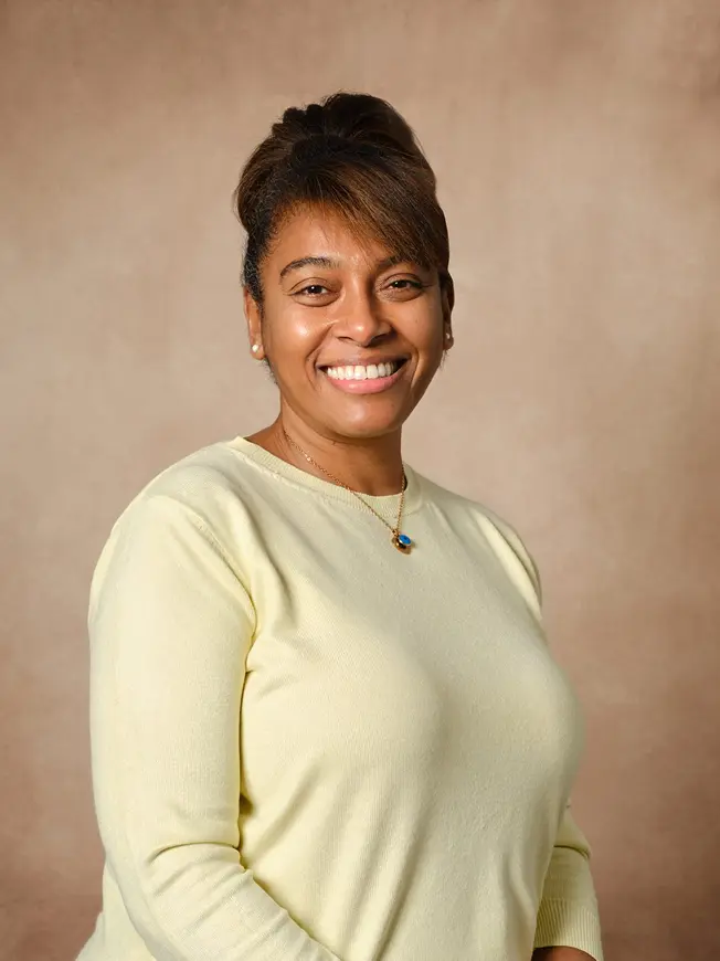 Smiling woman with light brown skin wearing a pale yellow sweater and a necklace against a beige background.