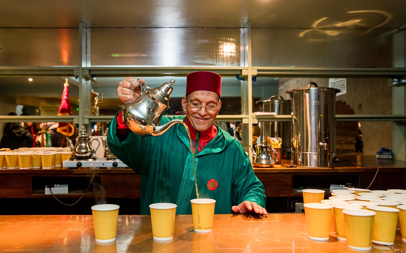Man in traditional attire pouring tea from a silver teapot into yellow cups on a counter.