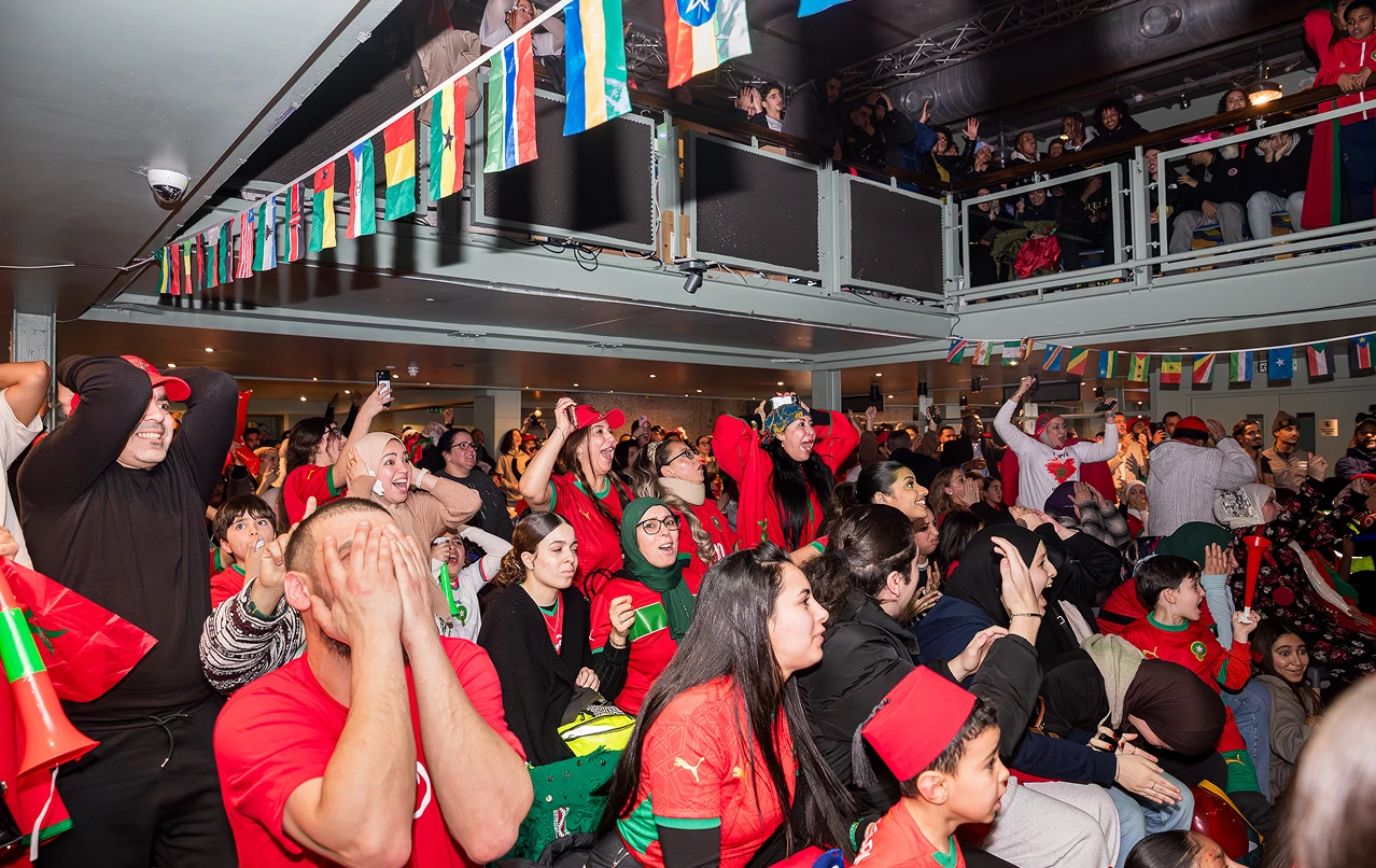 Crowd of excited people, many in red, watching an event indoors with international flags overhead.
