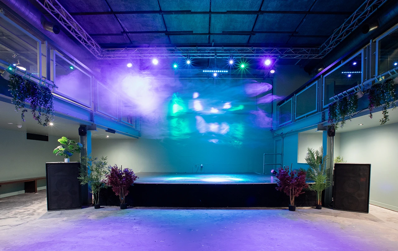 Empty indoor stage lit with colorful purple and blue lights, flanked by speakers and potted plants.