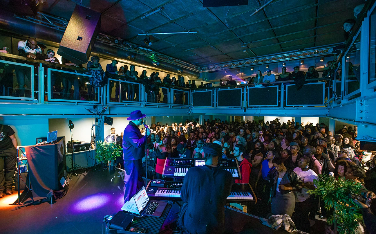 Musician in a hat performing on stage in front of a large crowd in a two-level venue.