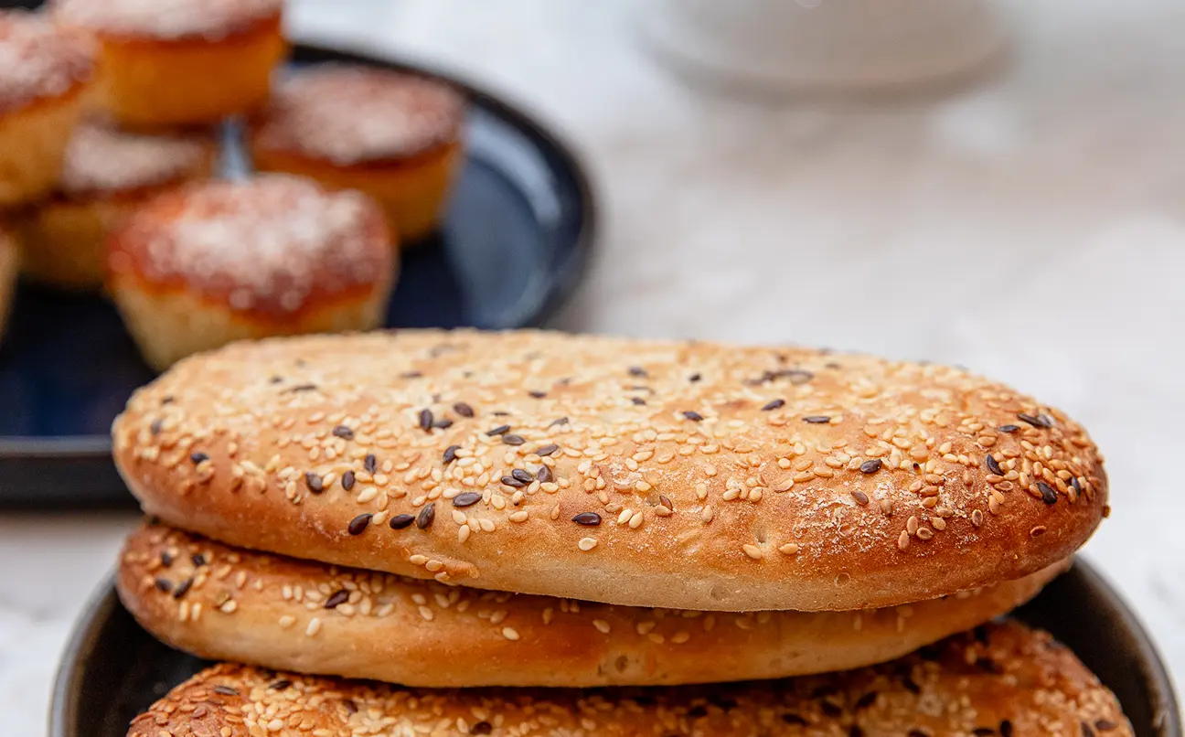 Oval sesame and seed bread loaves stacked in a bowl with small muffins in background.