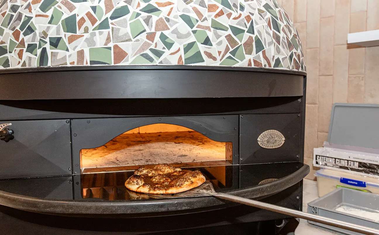 Wood-fired pizza oven with a pizza peel holding a partially baked pizza at the oven entrance.