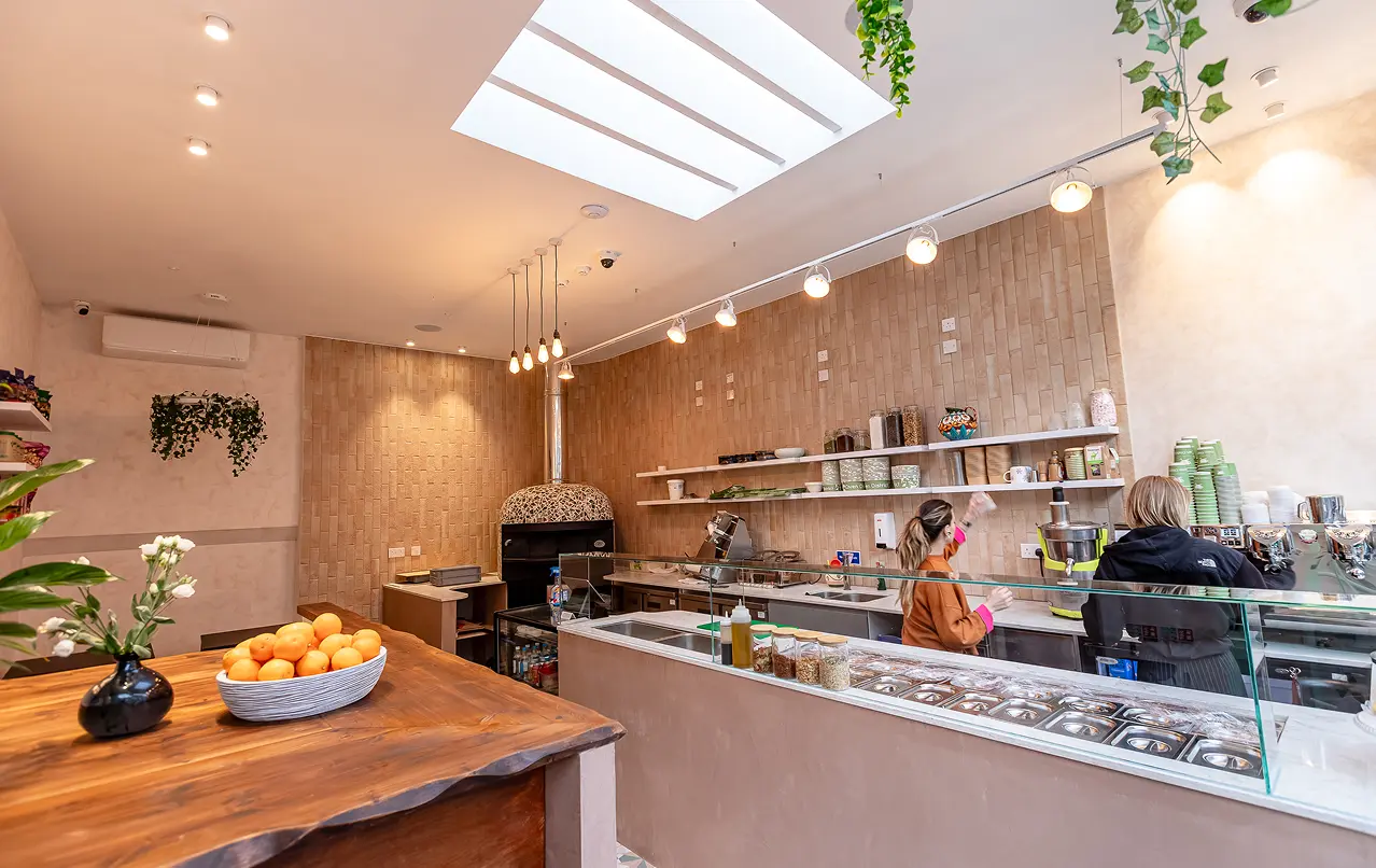 Bright modern cafe interior with wooden counter, fruit bowl, two staff working behind glass counter.