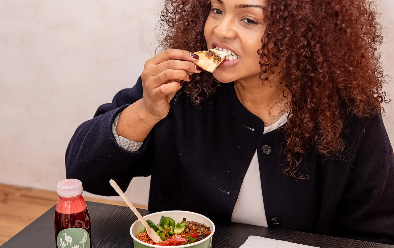 Woman with curly hair eating a slice of bread next to a bowl of food and a bottled drink on a table.