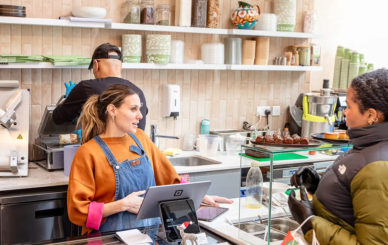 Barista in orange sweater and denim apron talking to a customer in a cafe with pastries on counter.