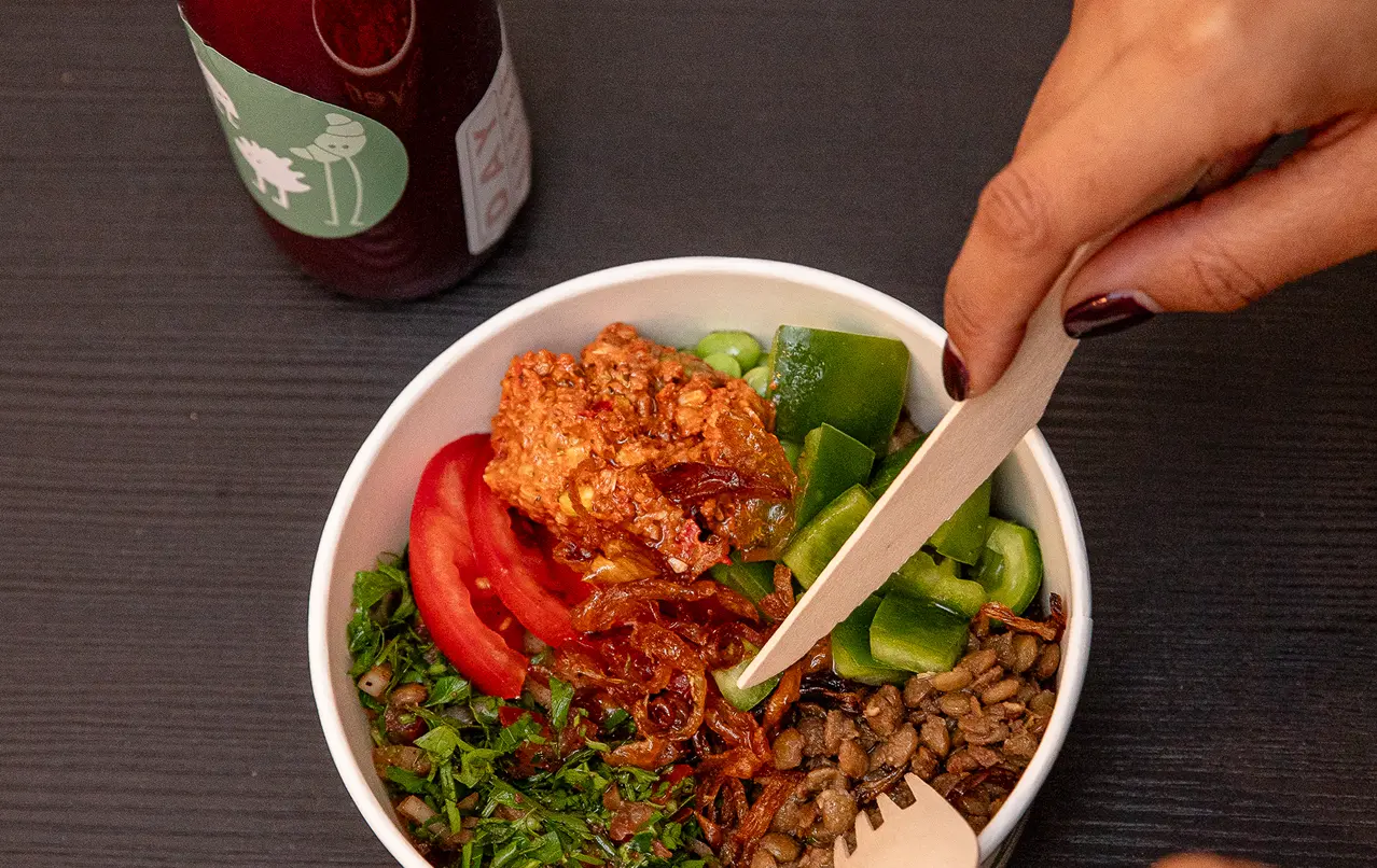 Hand holding a wooden fork and knife over a bowl with lentils, tomato slices, greens, and green peppers.