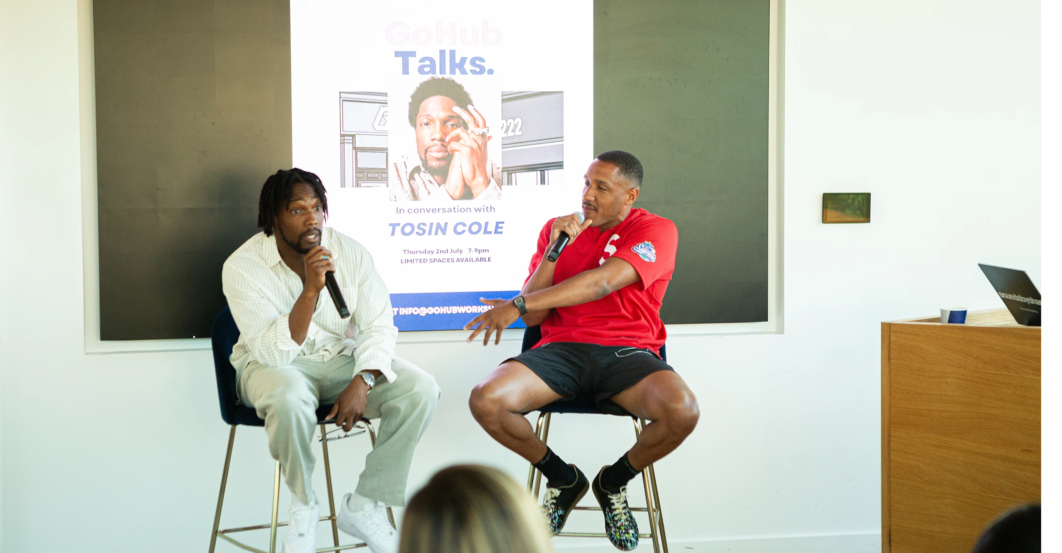 Two men sit on stools talking into microphones during a GoHub Talks event with Tosin Cole.