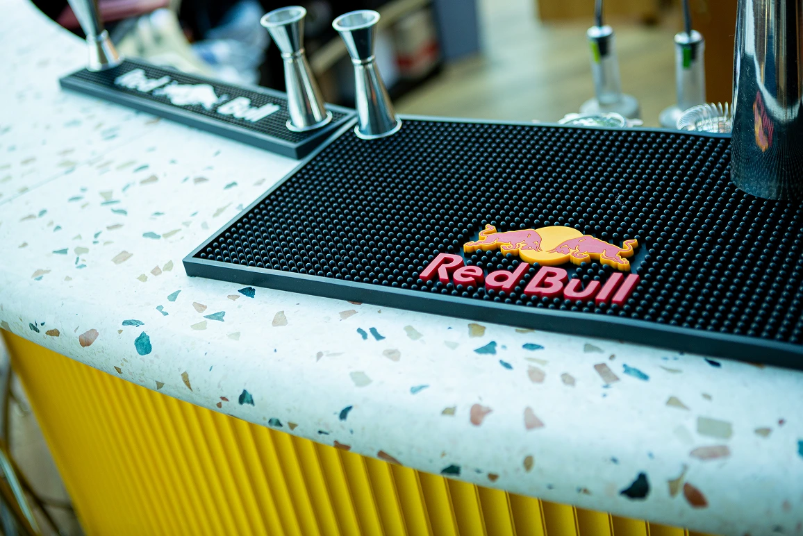 Close-up of Red Bull branded black bar mat with metal jiggers on a terrazzo countertop.