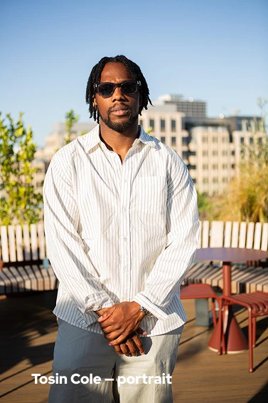 Man with sunglasses and dreadlocks wearing a white striped shirt outdoors with buildings in background.