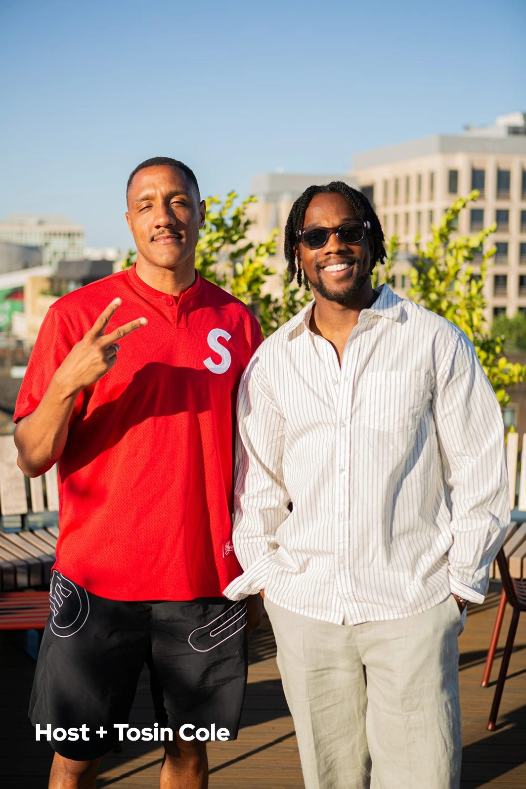 Two men outdoors; one wears red shirt showing peace sign, the other in striped shirt and sunglasses smiling.