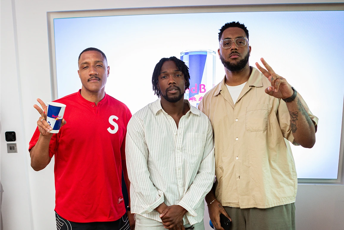Three men posing indoors: one in red holding a Red Bull cup, one in white, one in beige showing a peace sign.