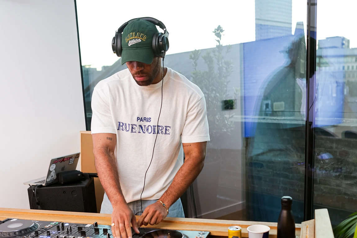 Man wearing green cap and headphones mixing music on DJ turntable indoors near window.