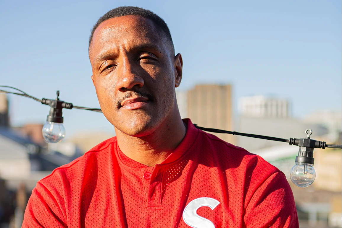 Man with short hair and mustache wearing a red shirt with letter S standing outdoors near string lights.
