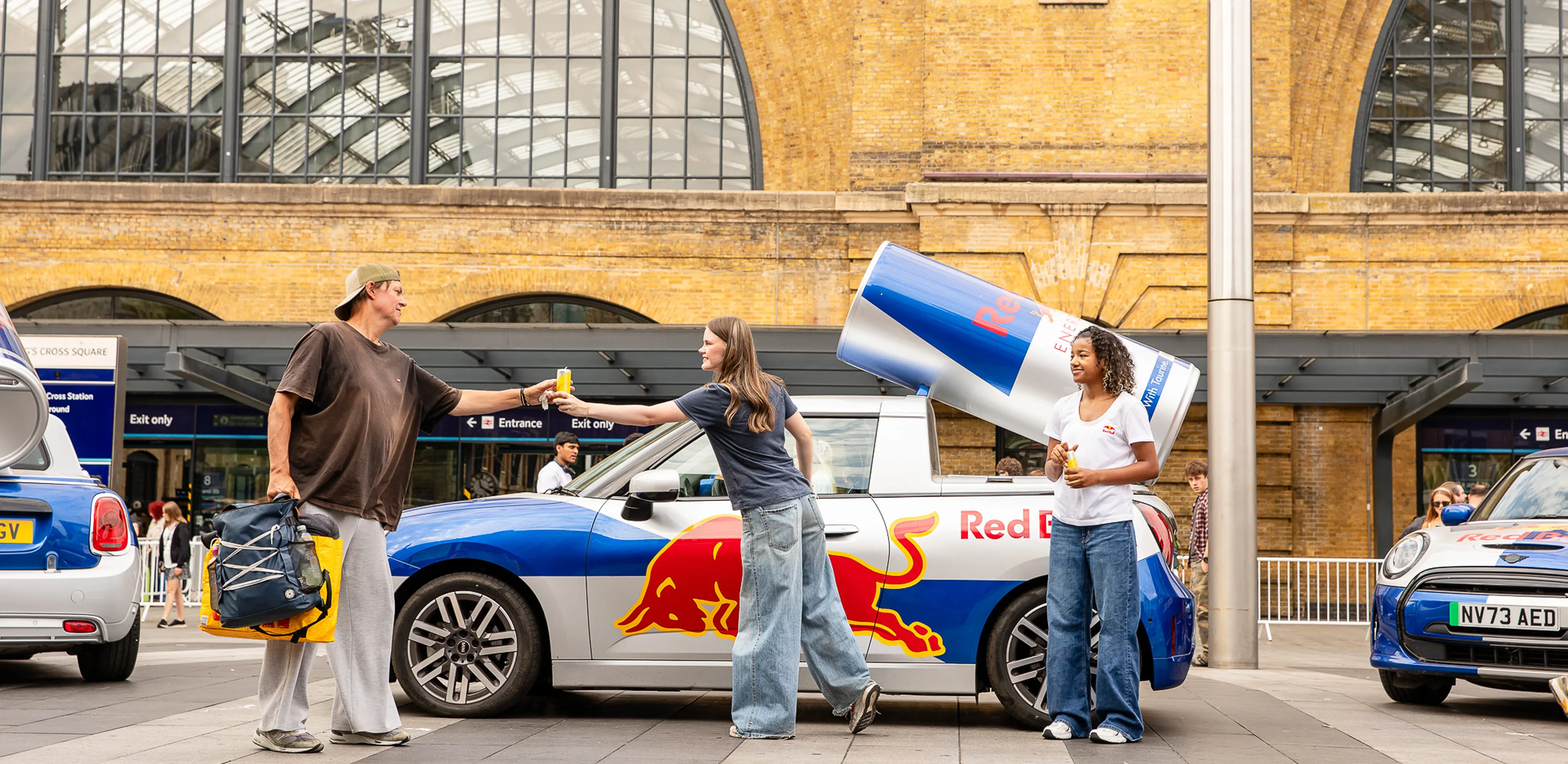 Two women hand out Red Bull cans near a branded Red Bull car with giant can sculpture on its roof.