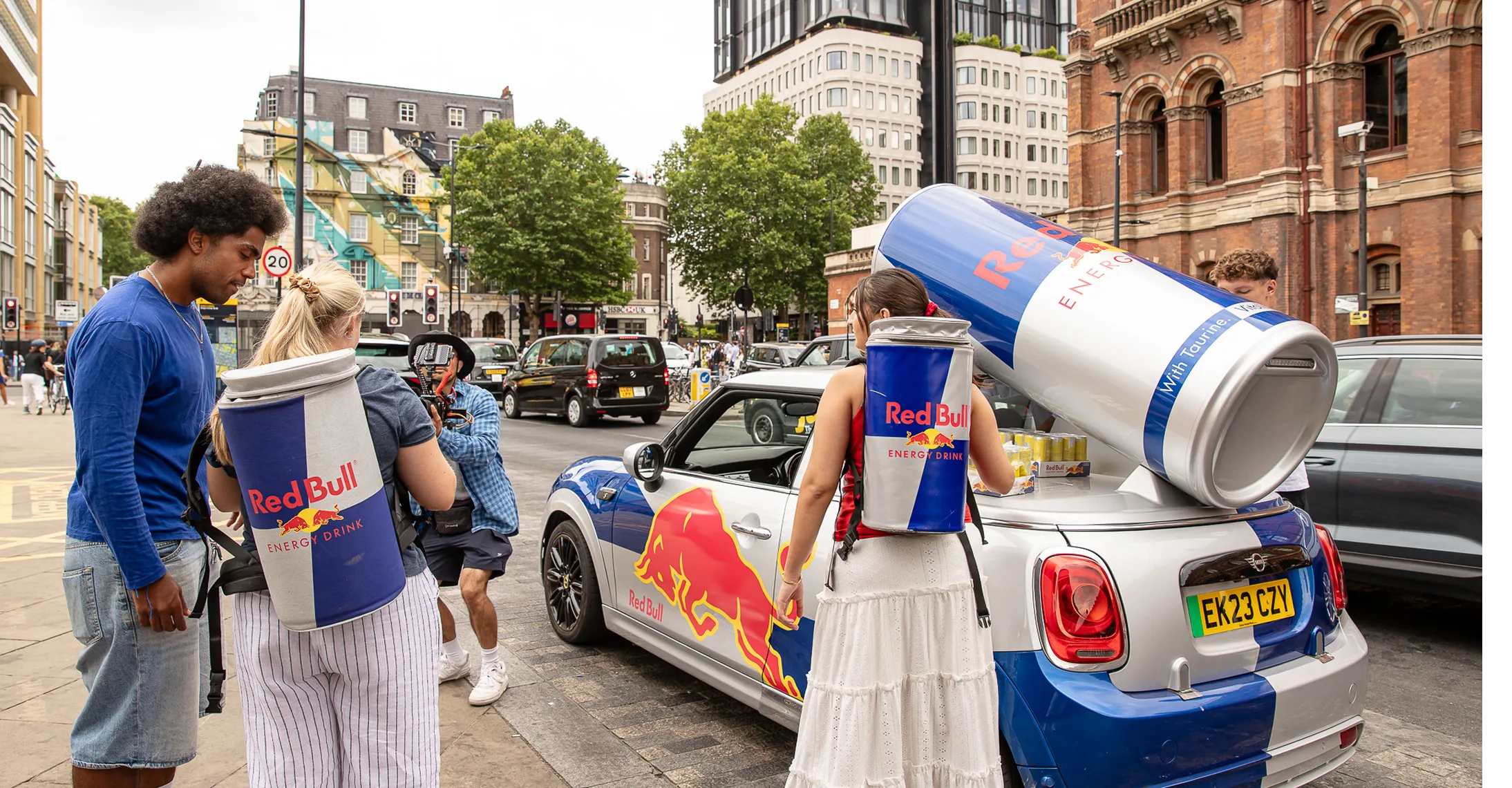 Two people wearing Red Bull cans backpacks stand near a Red Bull branded car with giant can on its roof.