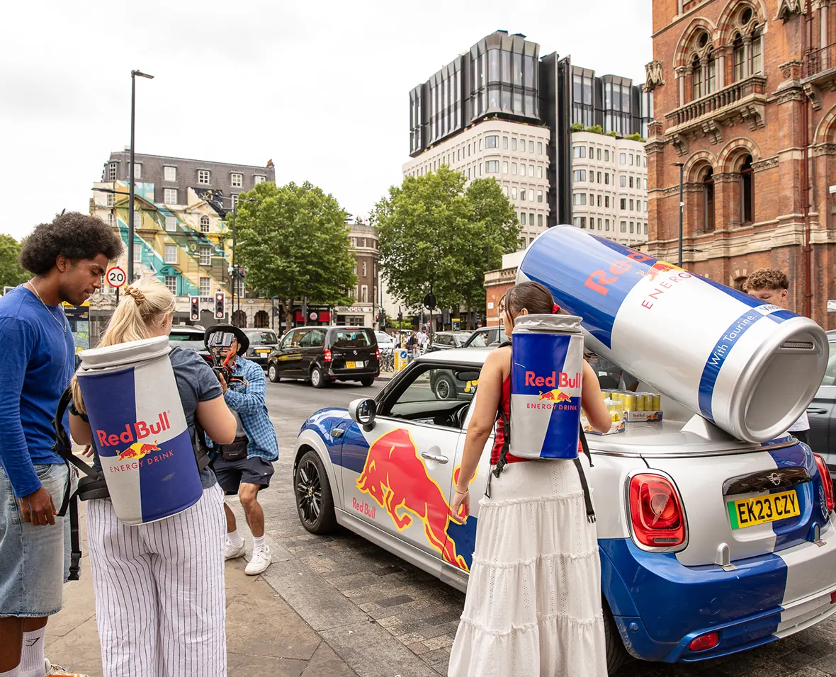 People with Red Bull can backpacks next to a branded Mini Cooper with a giant can on its trunk in a city street.