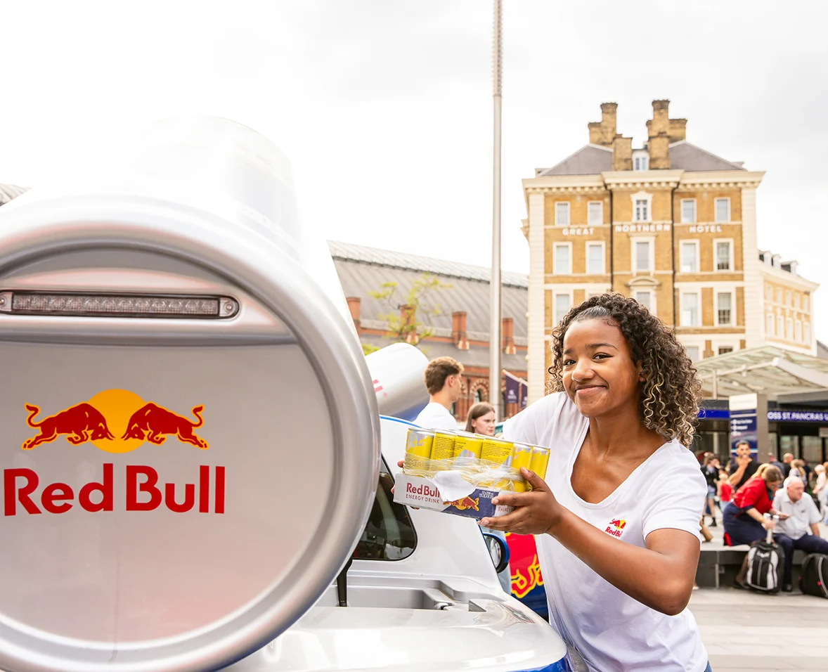 Smiling woman holding Red Bull energy drink cans near a branded vehicle in a busy urban plaza.