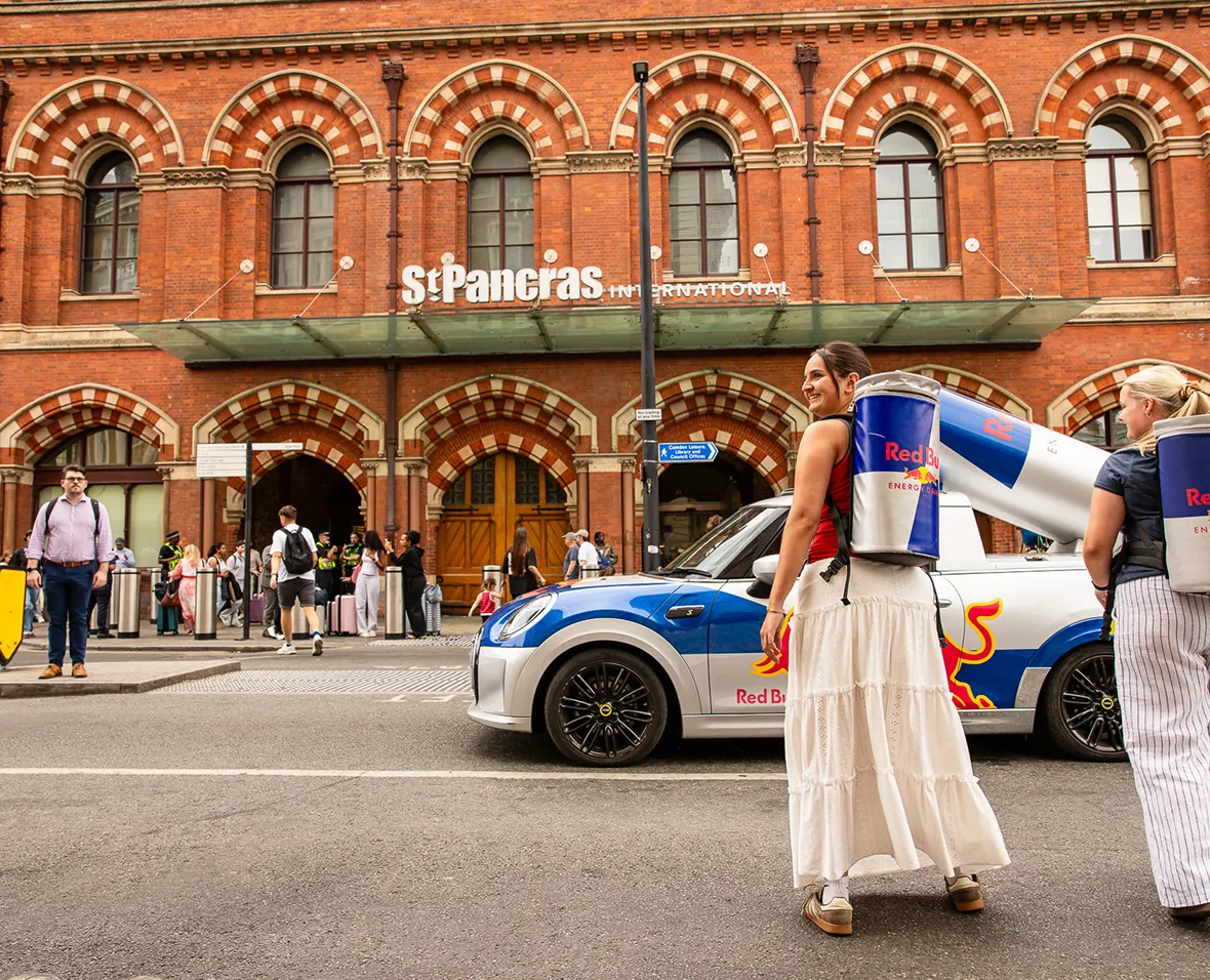 Two women wearing Red Bull can backpacks near a Red Bull car outside St Pancras International station.