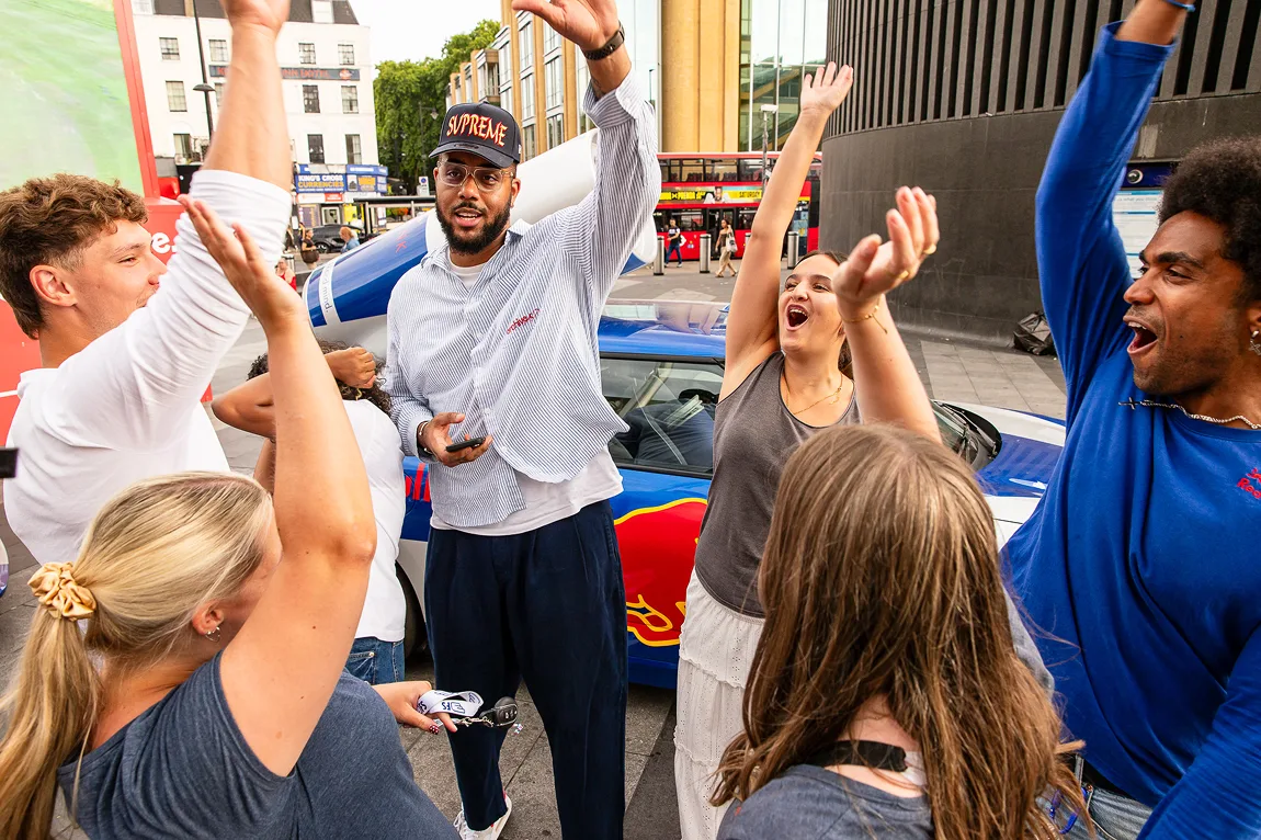 A diverse group of six people outdoors cheering and raising hands near a car with a Red Bull logo.