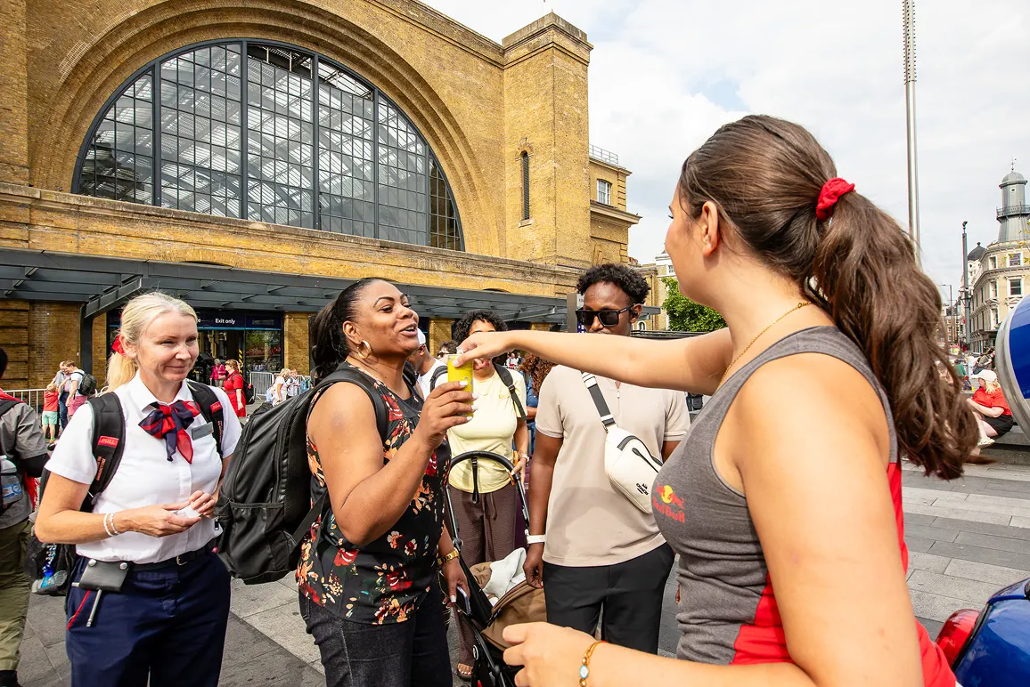 Group of people outside a train station, one woman handing a yellow object to another woman.