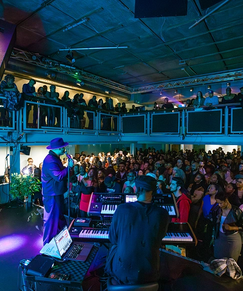 Musician in hat singing on stage with keyboard player in front of a large audience in an indoor venue.