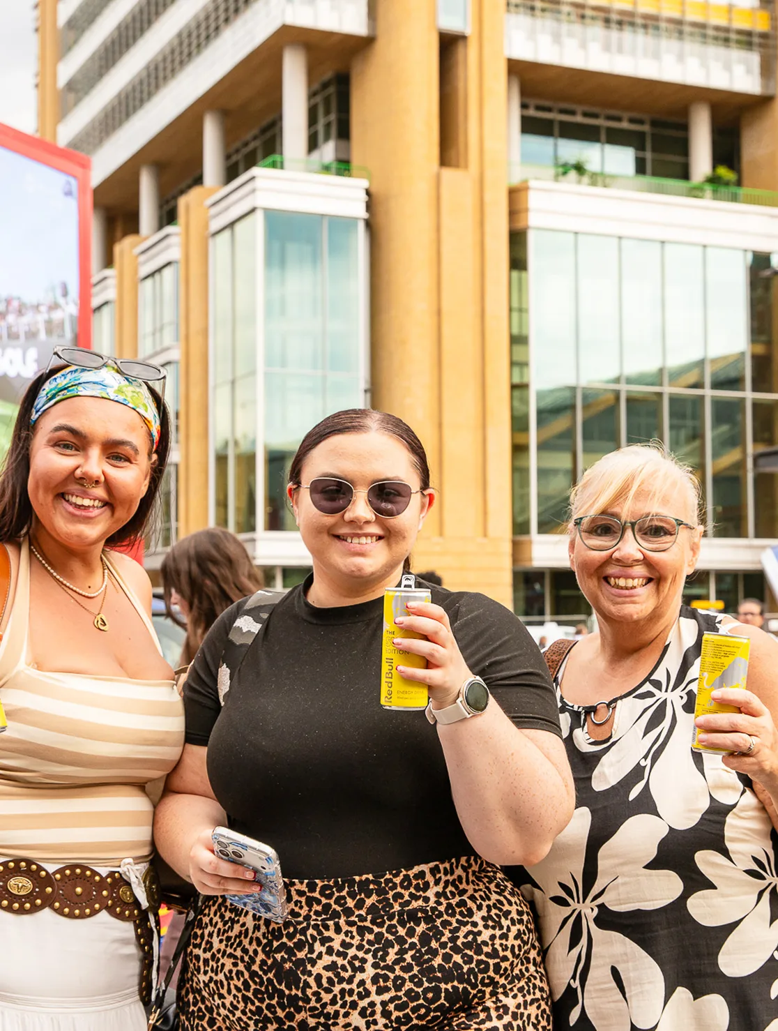 Three smiling women outdoors holding yellow cans of Red Bull energy drink in front of a modern building.