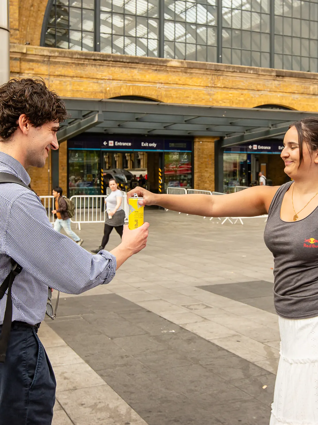 Woman in Red Bull tank top handing a can of Red Bull to a smiling man outside a train station.