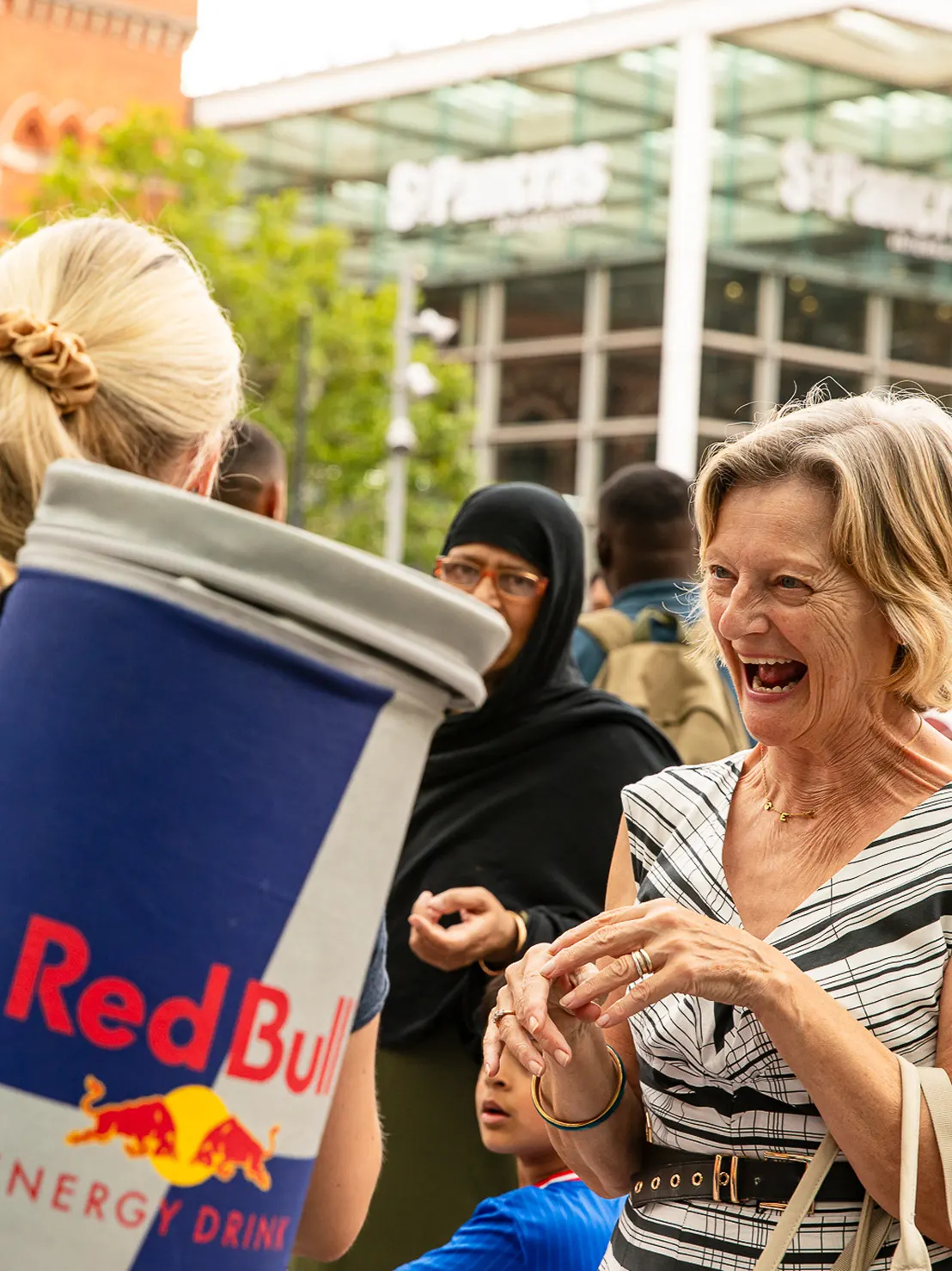 Smiling older woman engaging with person dressed as large Red Bull energy drink can in a busy outdoor area.