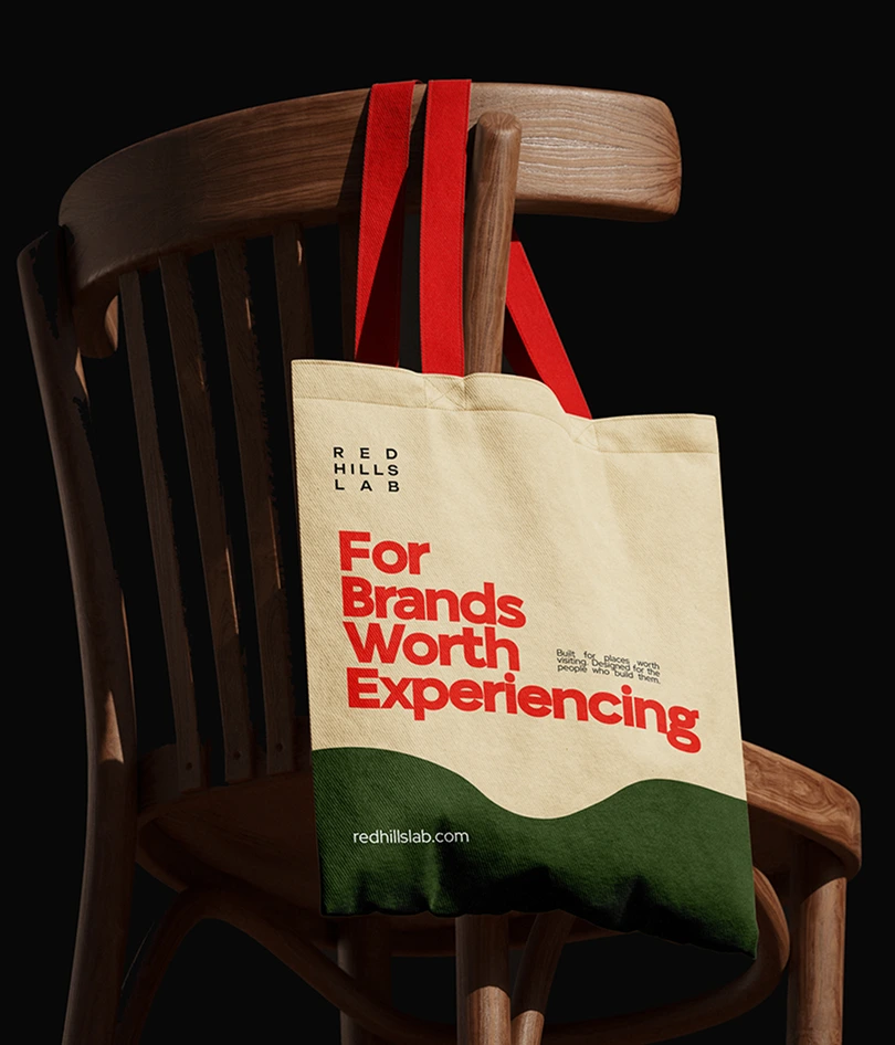 Canvas tote bag with red handles hanging on a wooden chair, text reads For Brands Worth Experiencing.