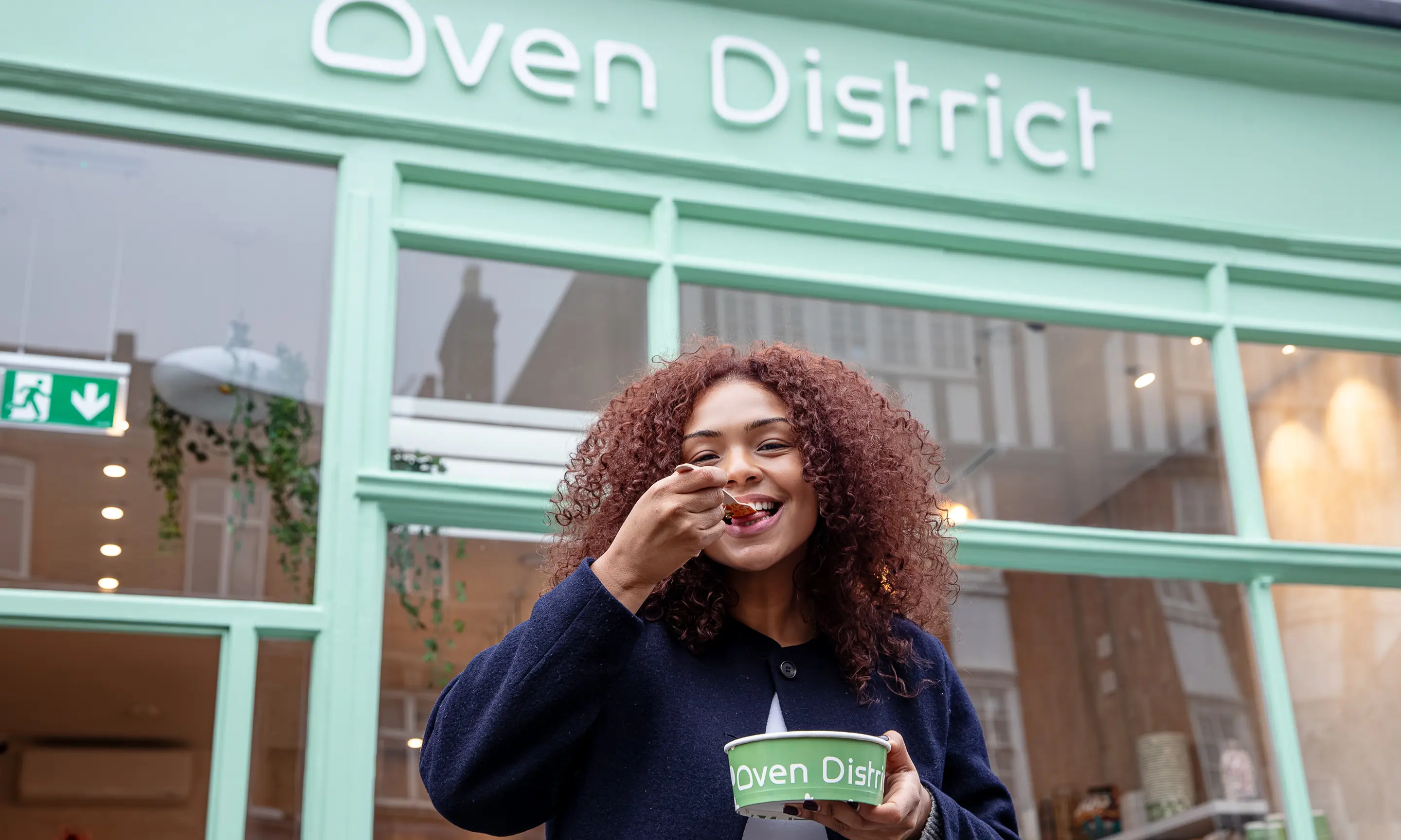 Smiling woman with curly hair eating food from a green bowl outside a mint green eatery.