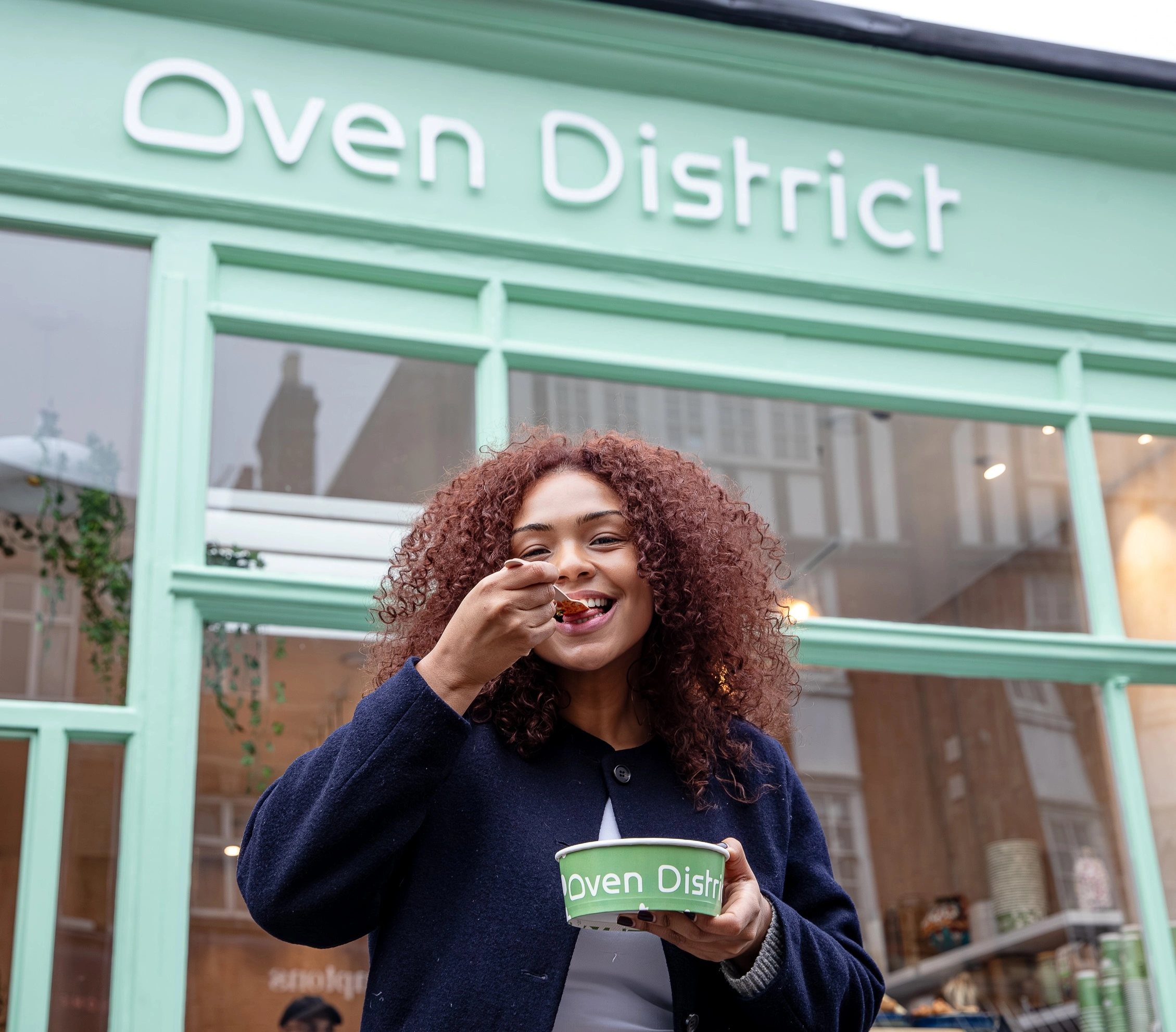 Smiling woman with curly hair eating food from a green Oven District bowl outside the shop.