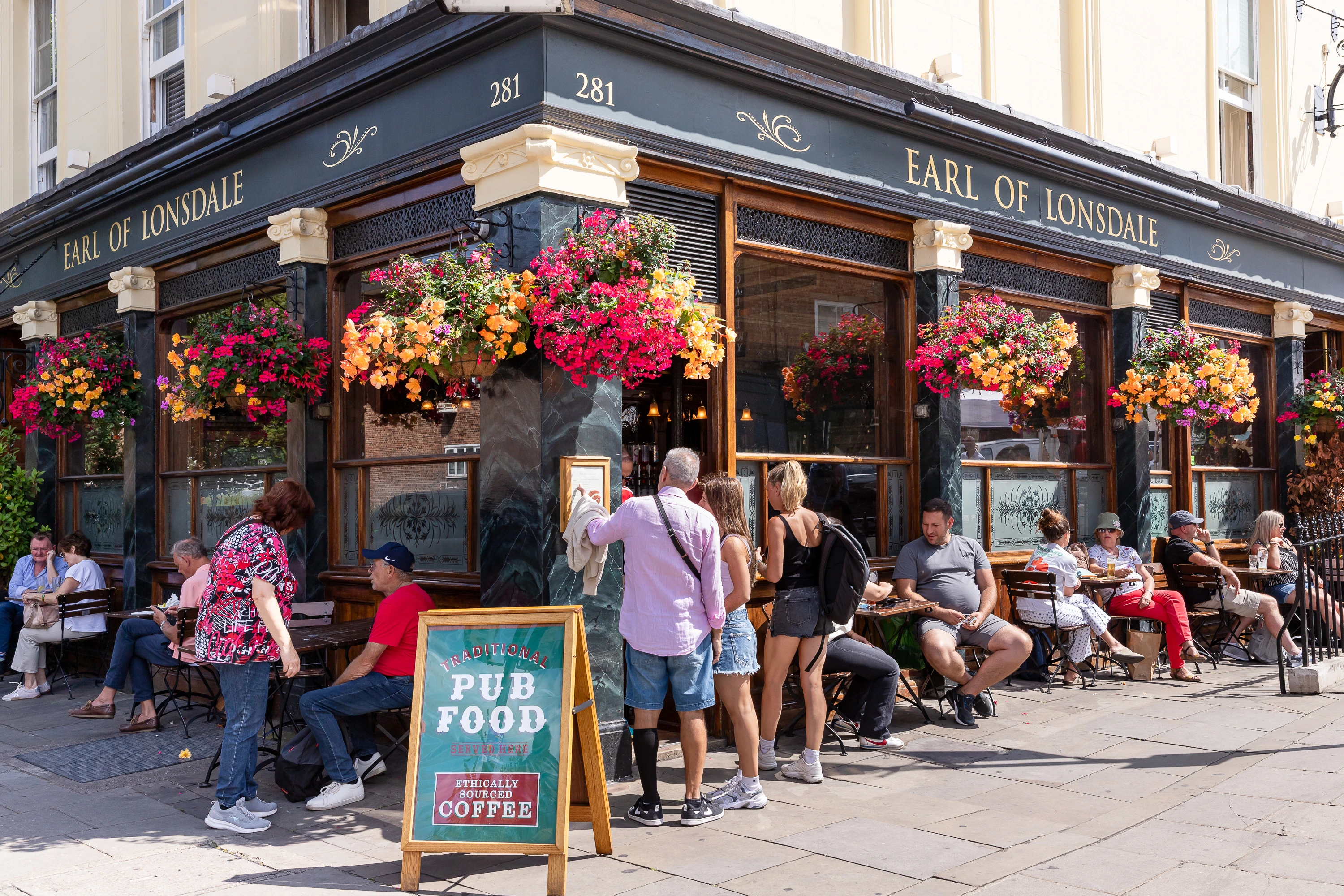 People outside Earl of Lonsdale pub with hanging flowers and a sign advertising traditional pub food and coffee.