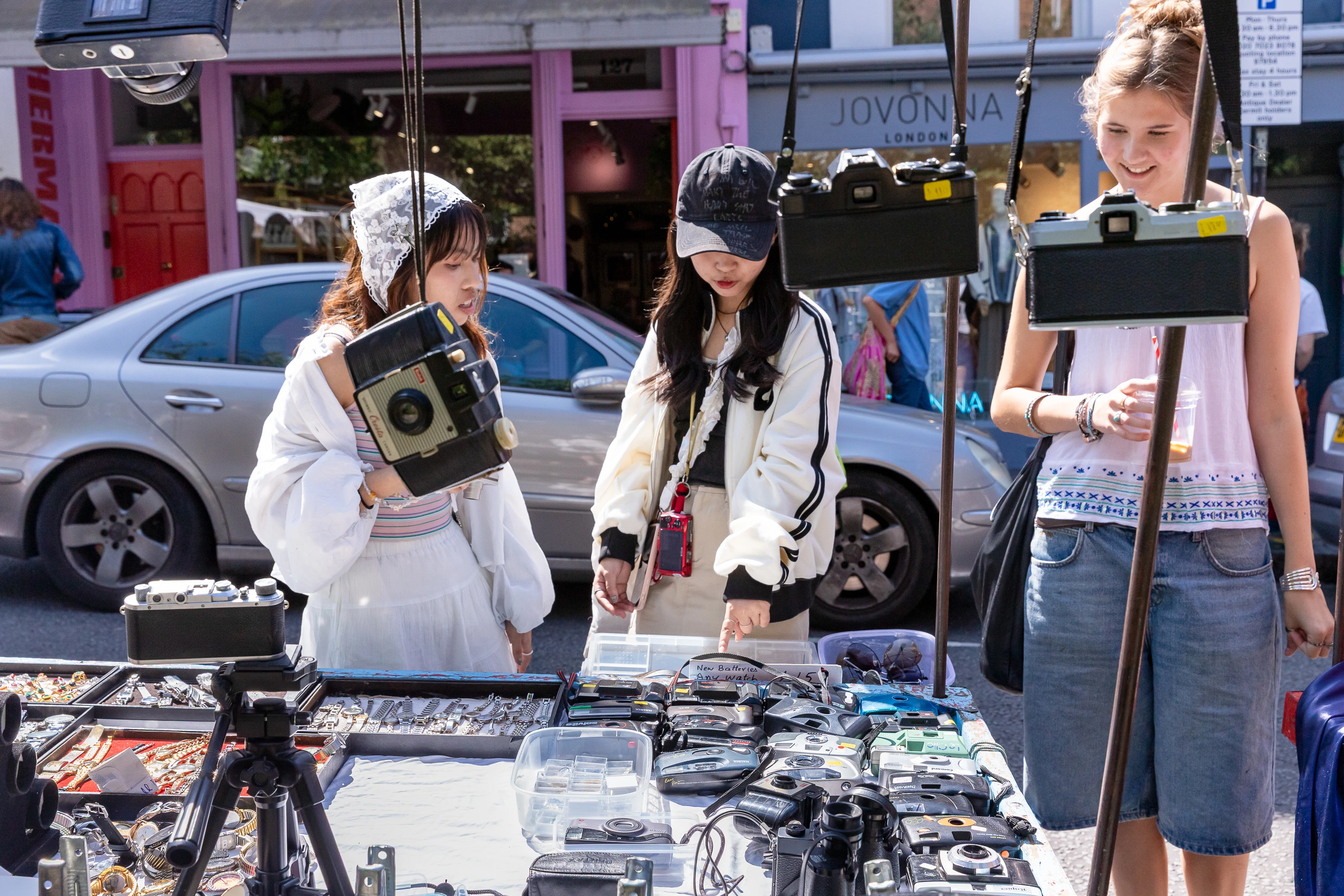 Three women browsing vintage cameras and watches at an outdoor market stall on a sunny day.