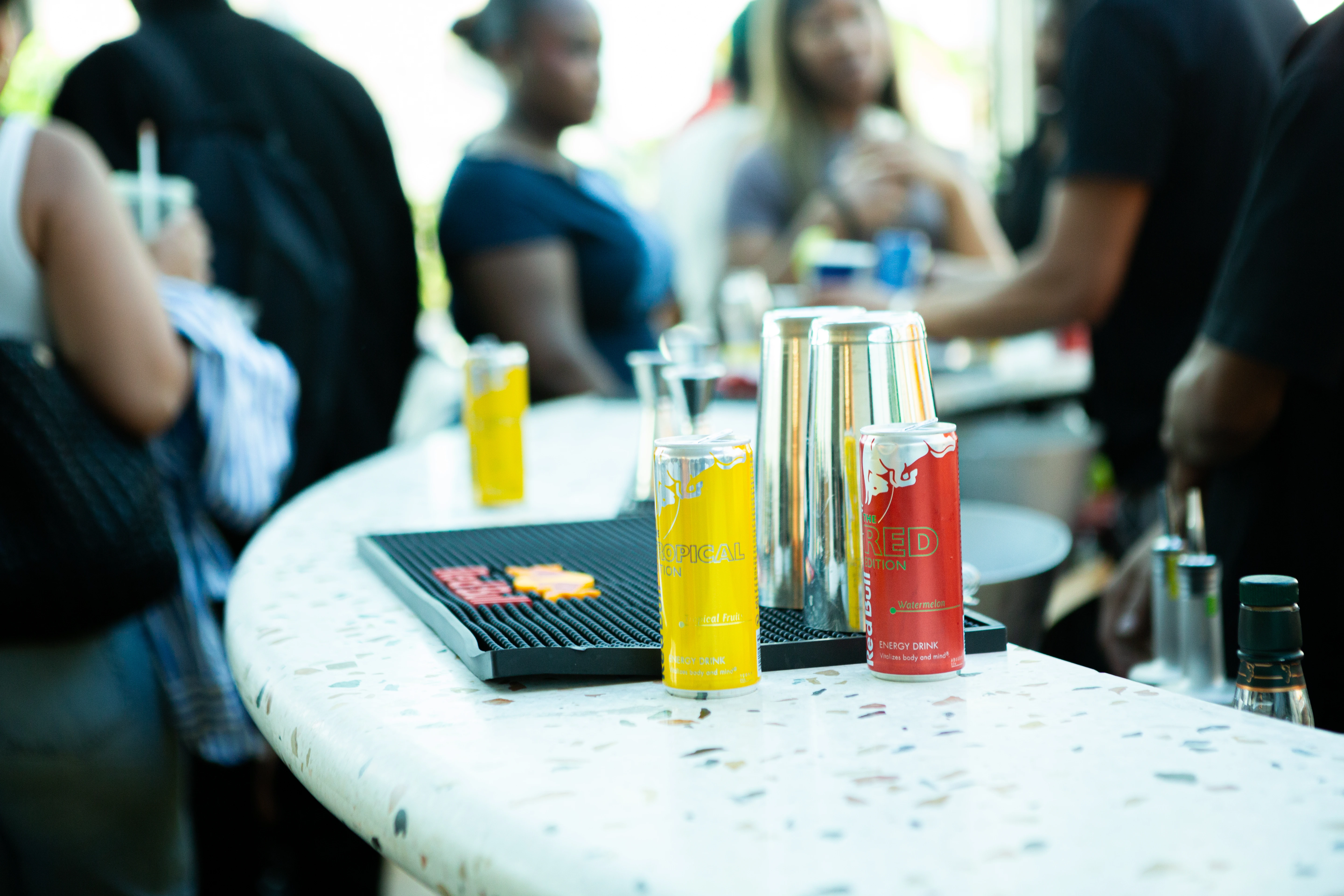 Red Bull Tropical and Red Edition cans on a bar counter with blurred people in background.