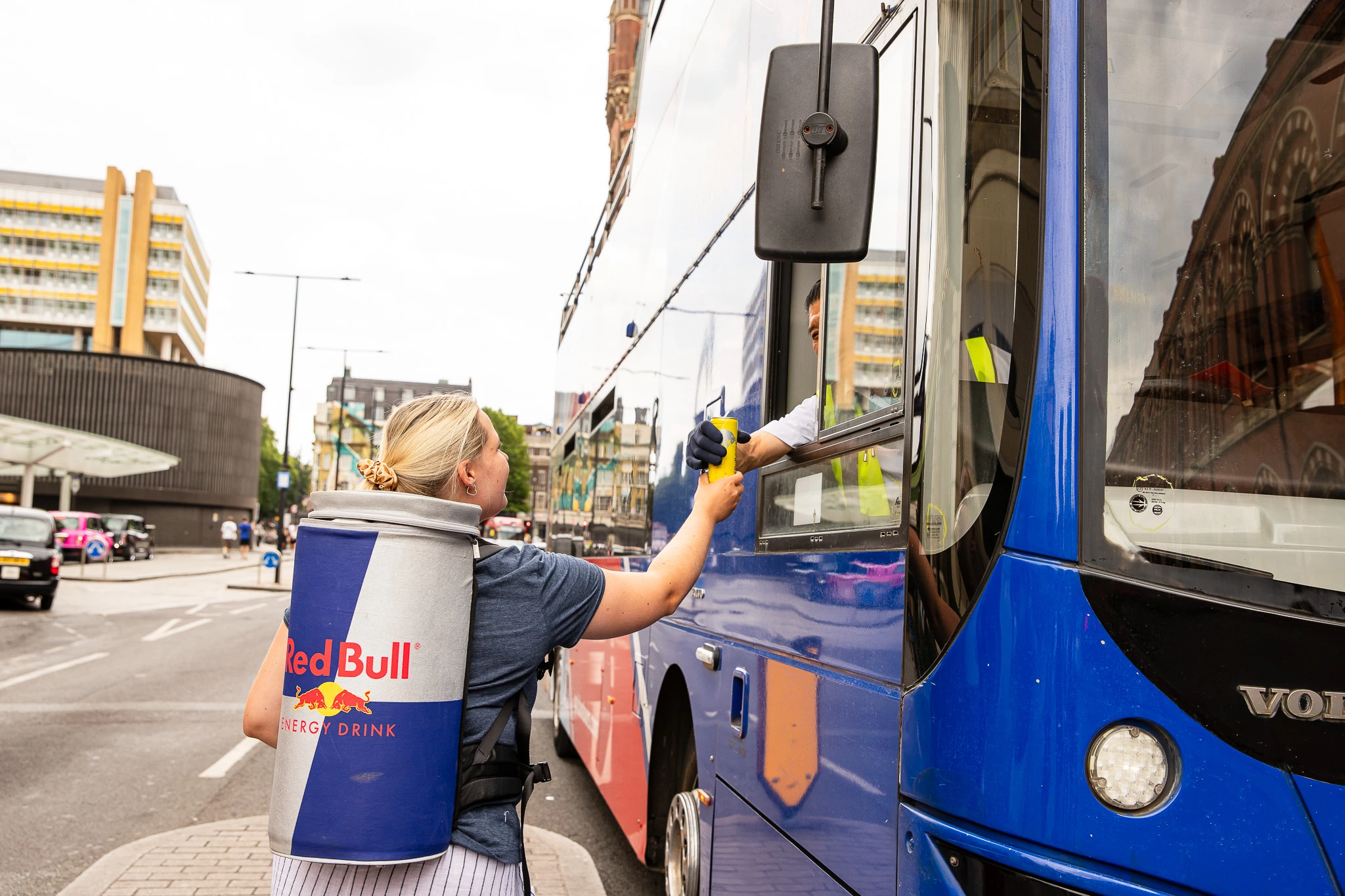 Person wearing large Red Bull can backpack handing a can to a bus driver through the window.