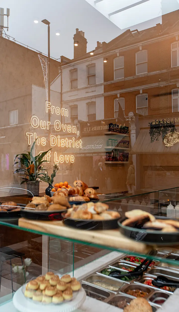 Bakery display with pastries and neon sign reading 'From Our Oven, The District of Love' reflecting buildings.