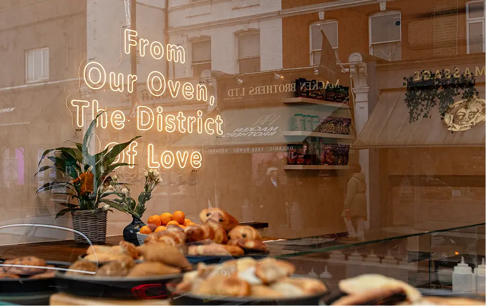 Bakery window with neon sign 'From Our Oven, The District of Love' and pastries displayed inside.