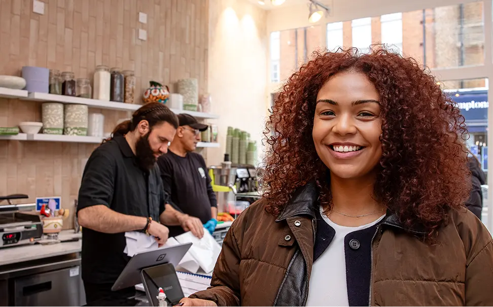 Smiling woman with curly hair in brown jacket stands in cafe with two men working behind counter.