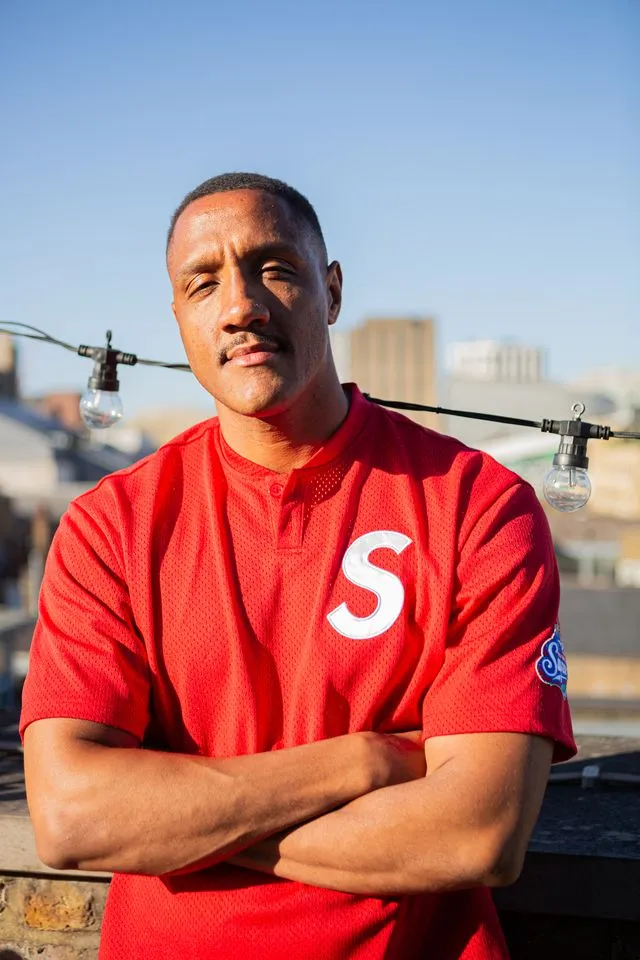 Man with short hair and mustache in a red shirt with S logo, standing with arms crossed outdoors.