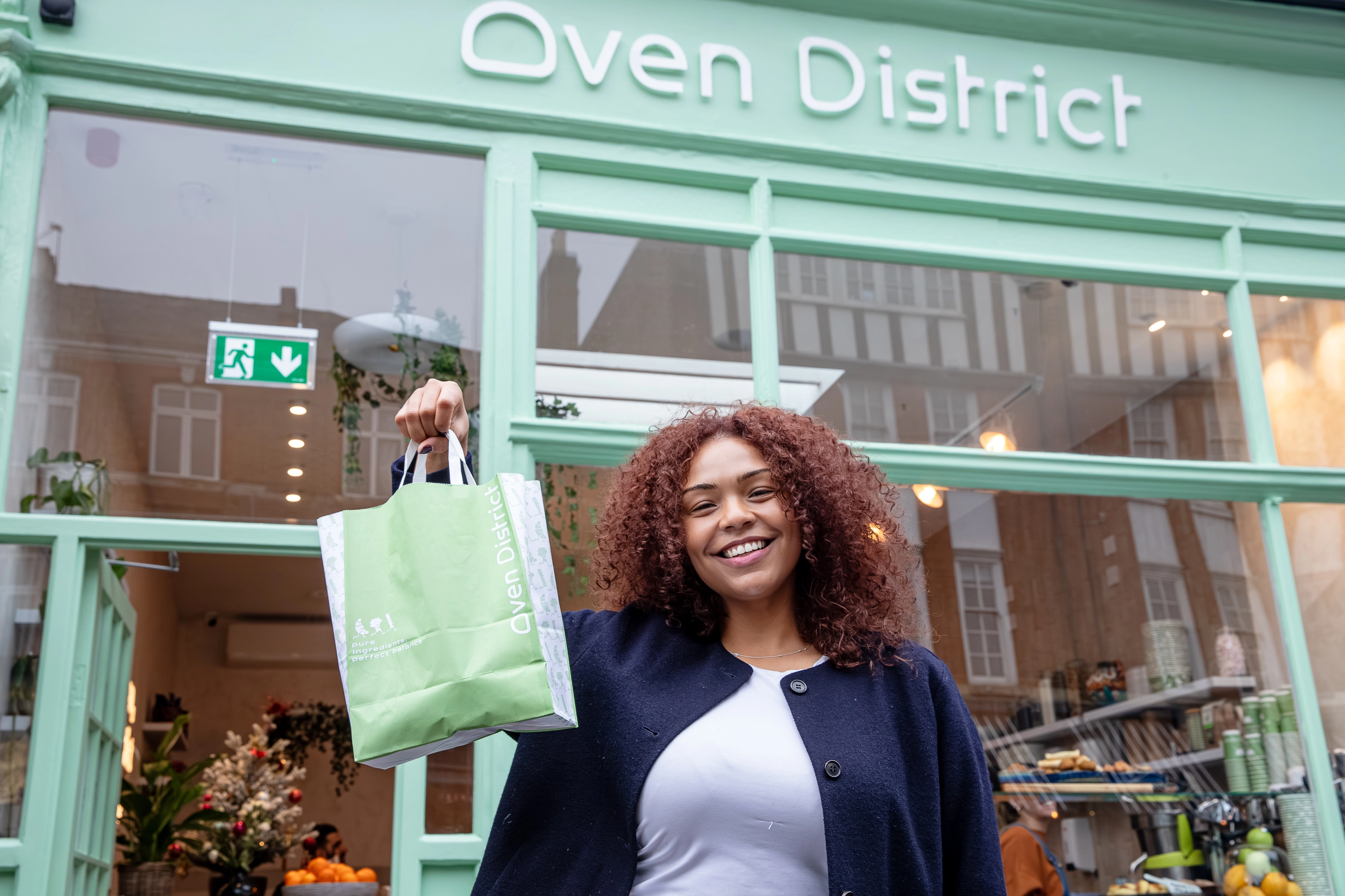Smiling woman holding a green Oven District shopping bag outside a mint green storefront.