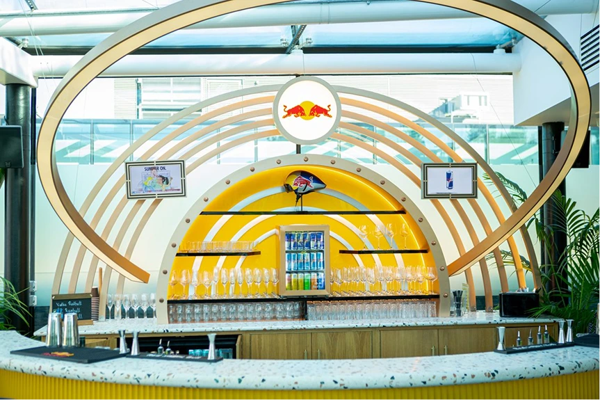 Bright modern bar with Red Bull logo, glass shelves, multiple glasses, and drink fridge in center.