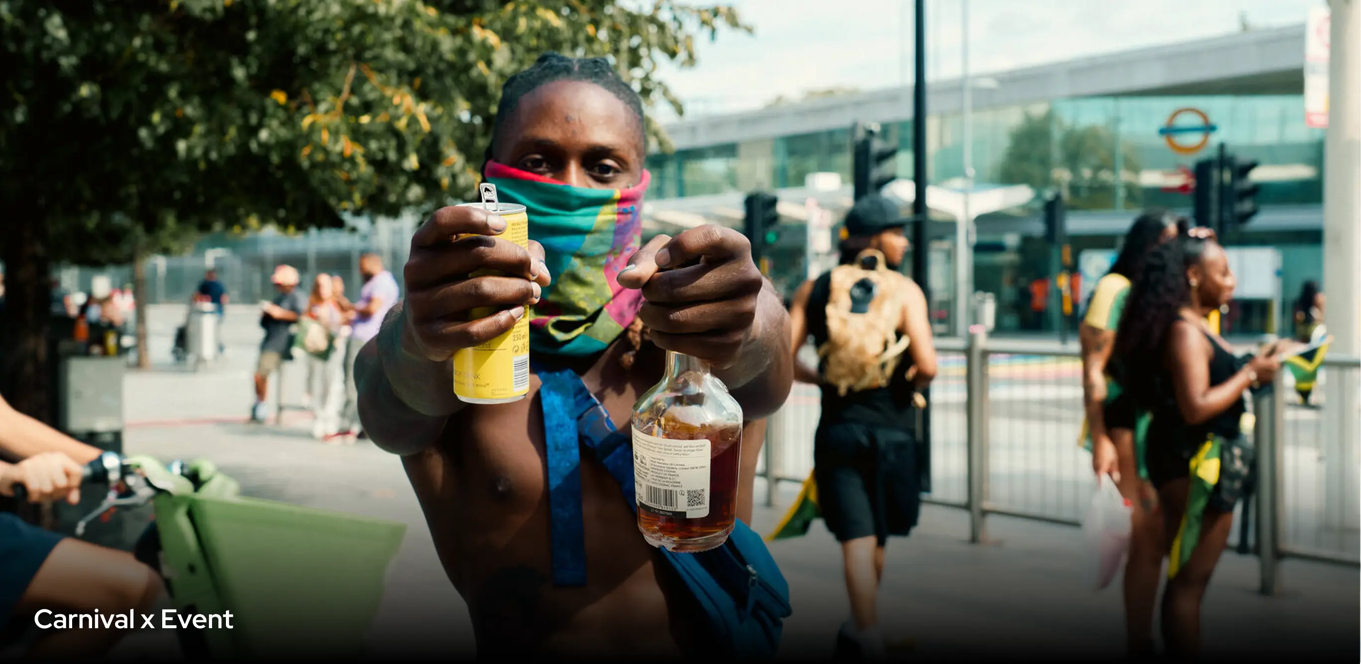 Shirtless man wearing a colorful face covering holds a can and a bottle of liquor toward the camera at a carnival event outdoors with people and urban surroundings in the background.