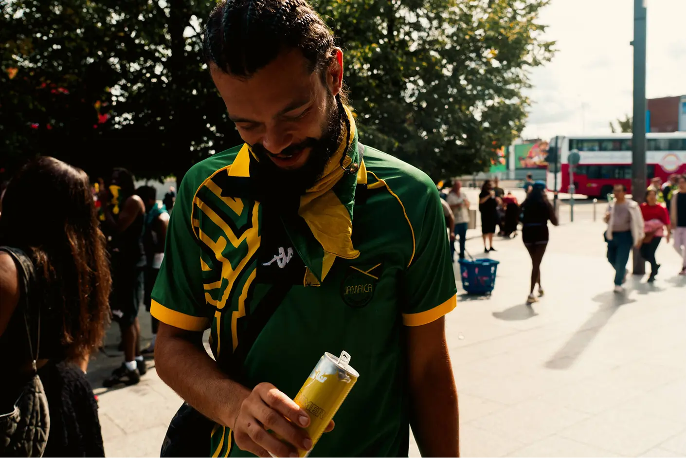 Man wearing a green and yellow Jamaica sports jersey and a matching scarf around his neck, holding a yellow and white can while looking down and smiling, with people and trees in the outdoor background.