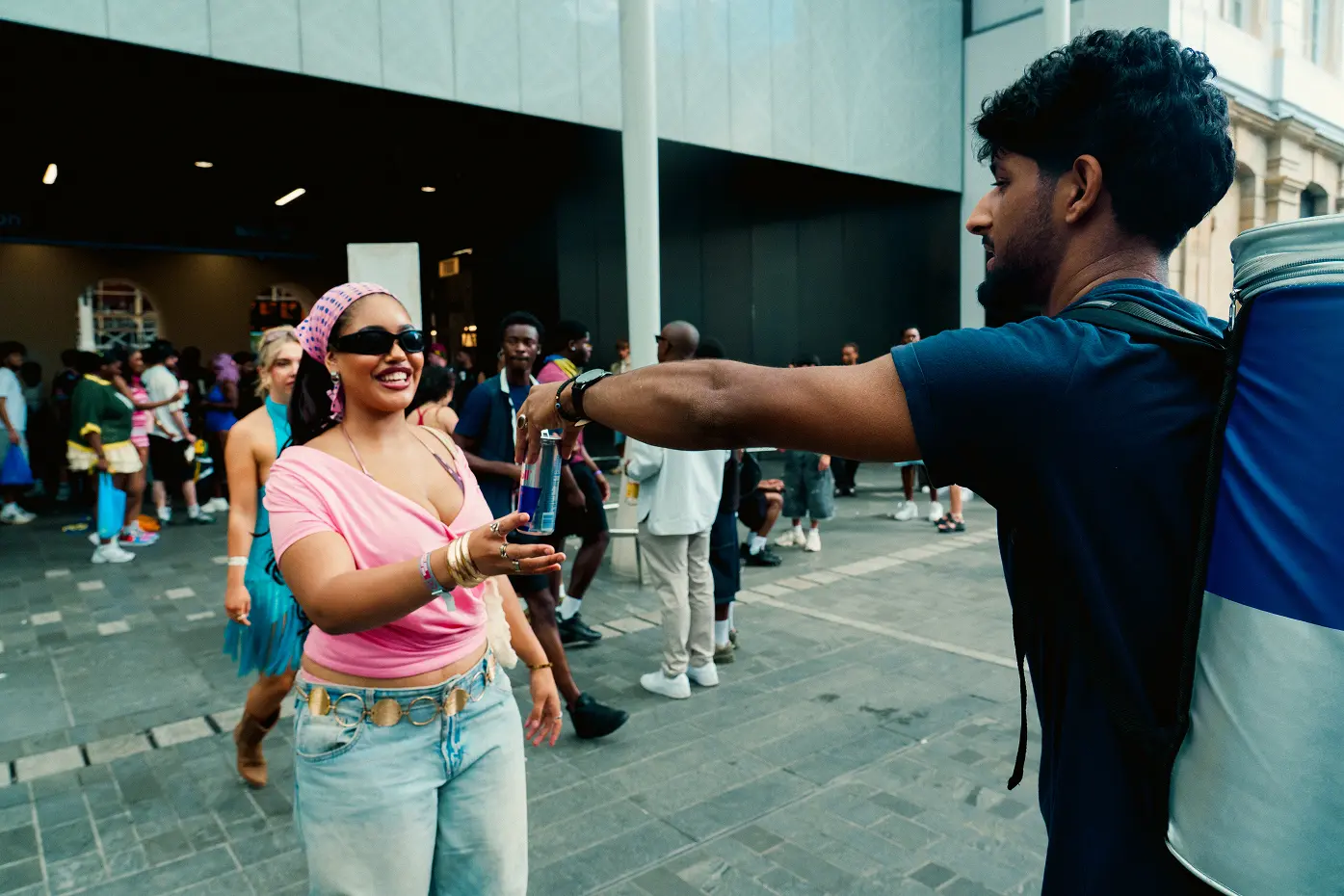 A man with a backpack cooler hands a can of Red Bull to a smiling woman wearing sunglasses, a pink top, and a pink headscarf in a busy outdoor urban area with people walking around.